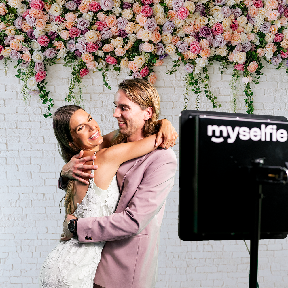 Brudpar kramas framför en blomstervägg bredvid Myselfie fotobås på bröllopet