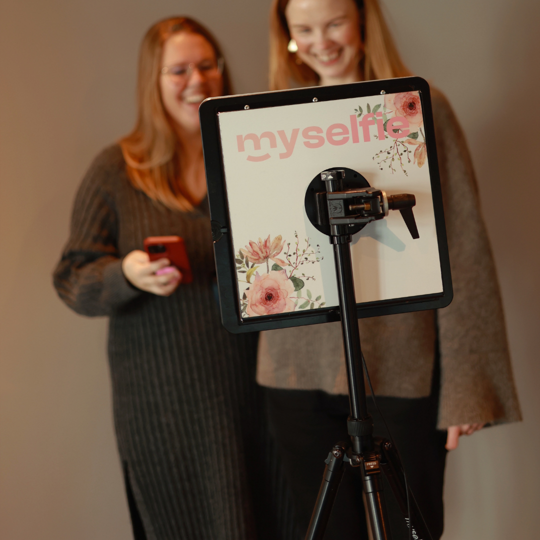 Två kvinnor tittar på en Myselfie fotobås med blommig ram